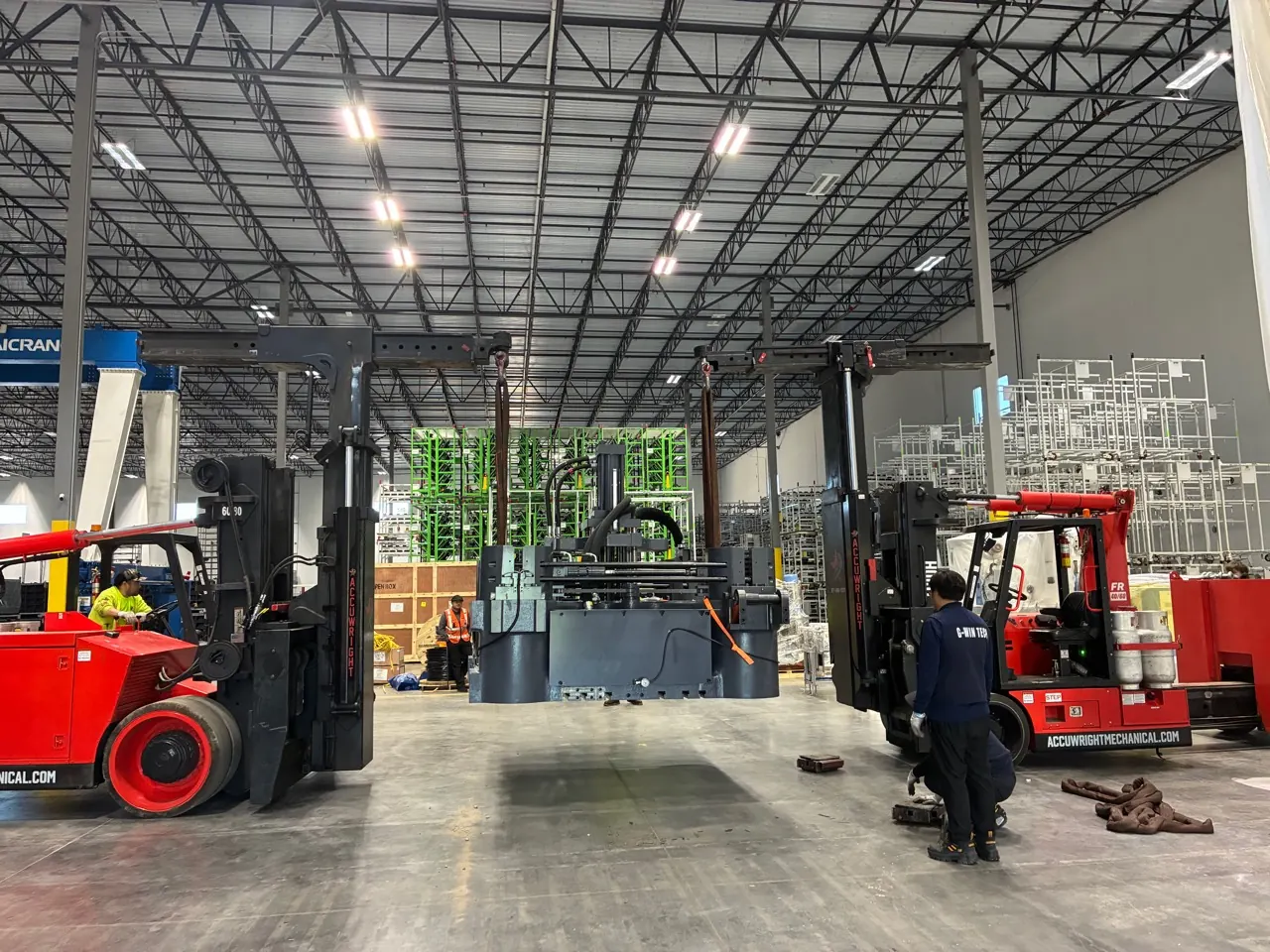 Two Accuwright heavy forklifts with boom attachments tandem-lifting a large machine base inside a plant