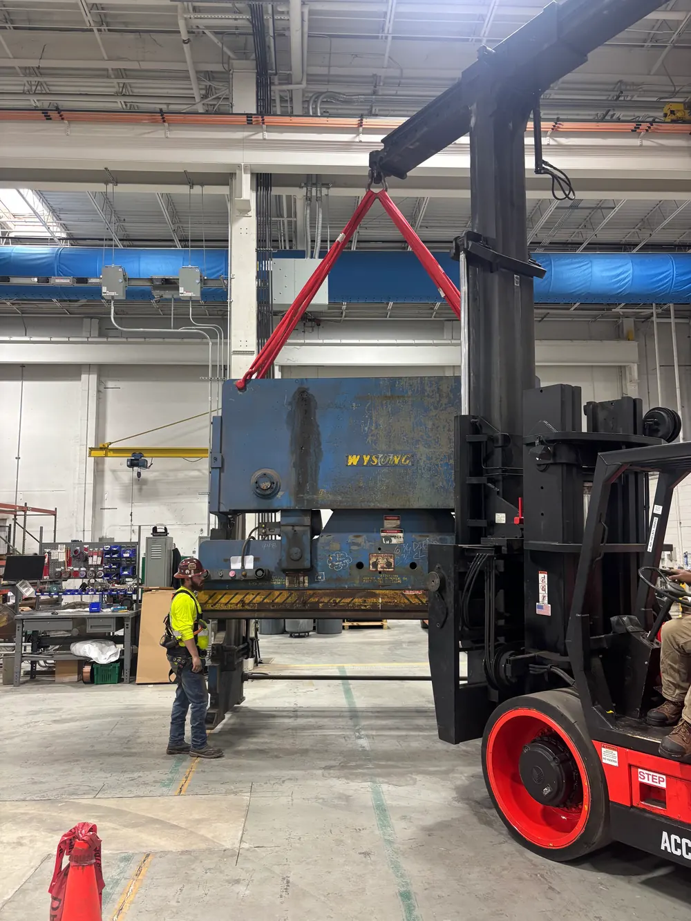A heavy forklift lifting a large press inside a plant, with a worker alongside for scale