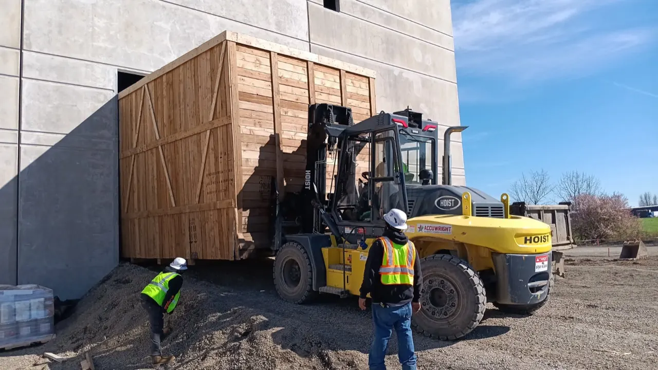 An Accuwright heavy forklift moving a large wooden crate through a plant wall opening