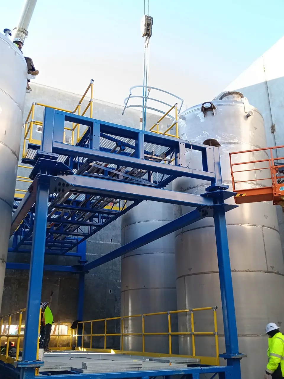 Stainless vessels and blue access platforms under active installation with workers and an overhead lift