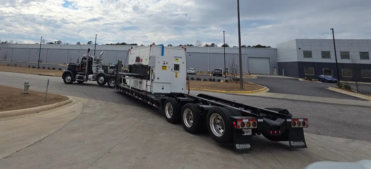 A lowboy trailer transporting a large machine through an industrial park
