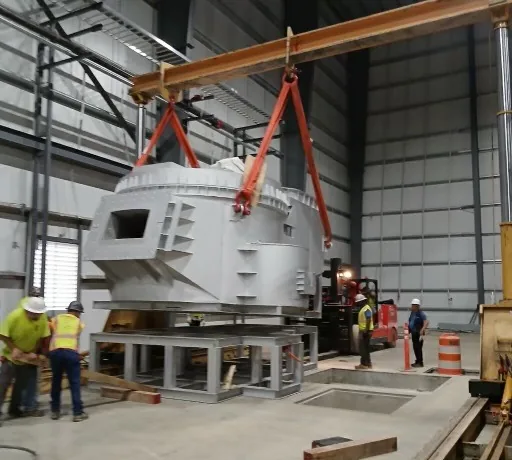 A large white machine component suspended from a lifting beam on red slings while Accuwright crew members spot the lift