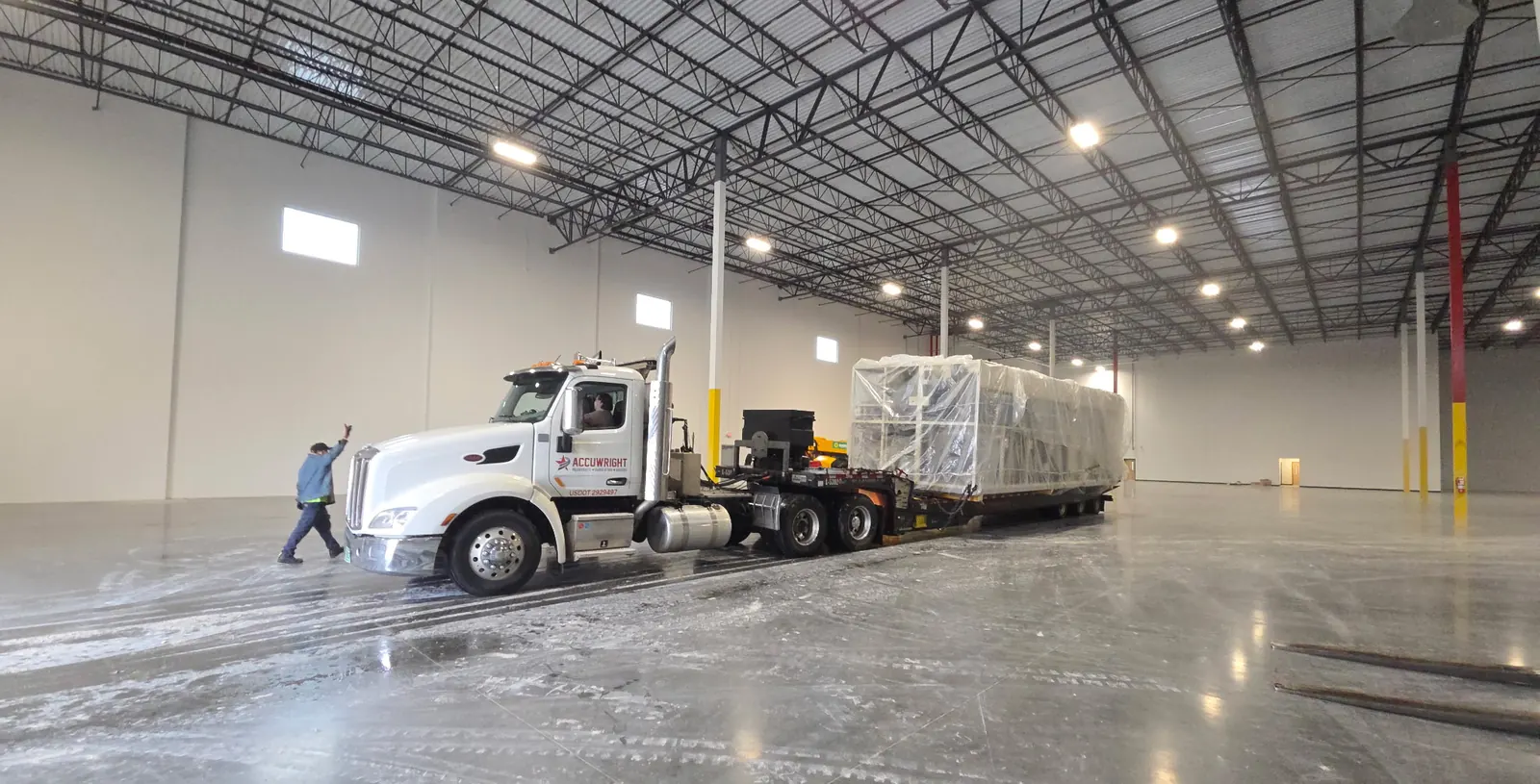 An Accuwright tractor and lowboy delivering a wrapped machine inside a large clean warehouse