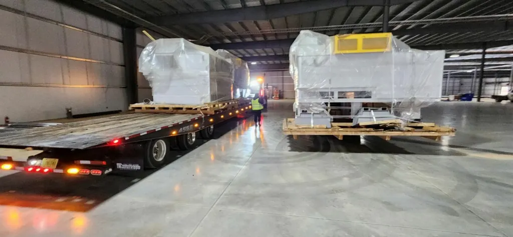 Wrapped machines staged on skids on a warehouse floor beside a flatbed trailer
