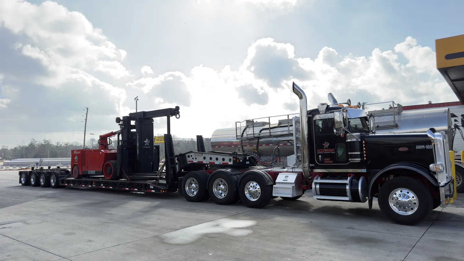 An Accuwright tractor and lowboy hauling a red Versa-Lift heavy forklift