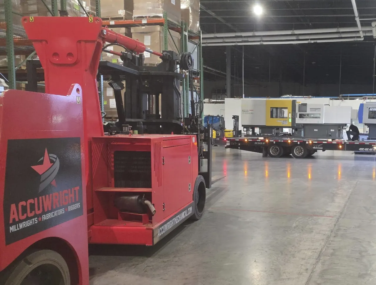 A branded red Accuwright machinery mover in the foreground as a flatbed trailer delivers machines inside a warehouse
