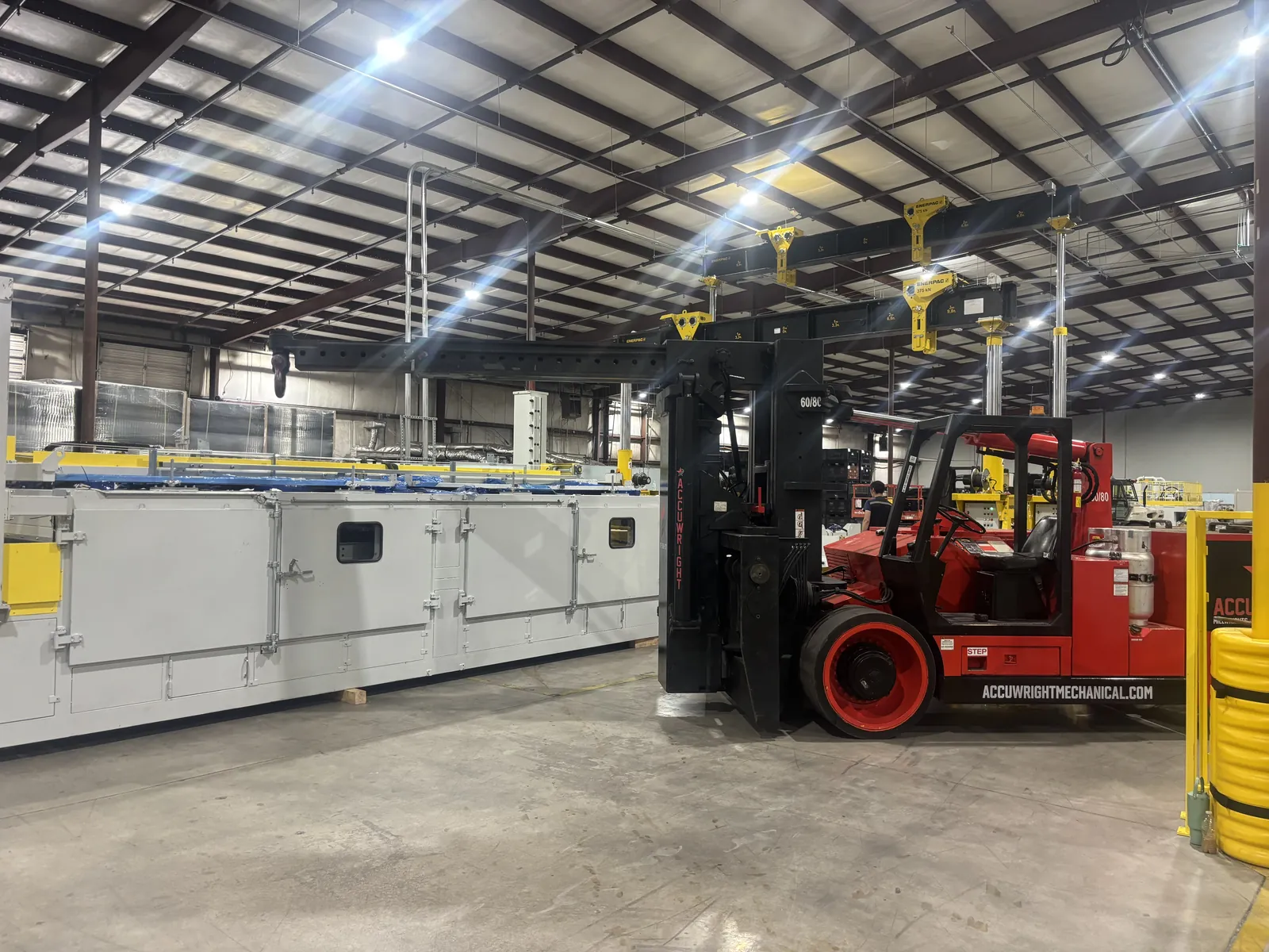 An Accuwright heavy forklift beside a long white machine line beneath a yellow hydraulic gantry system inside a plant