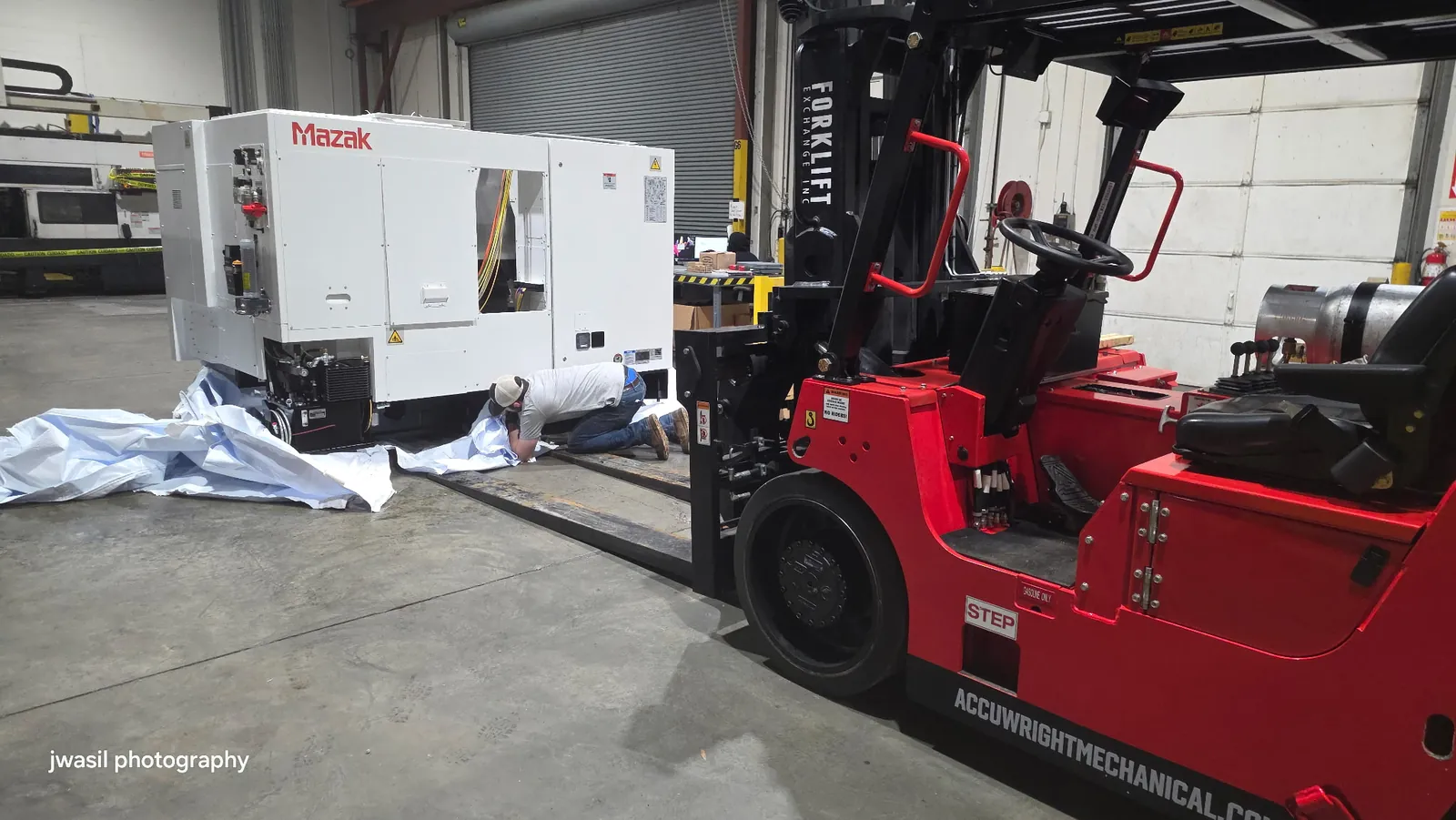 An Accuwright forklift setting a new white CNC machine while a worker kneels placing protective sheeting
