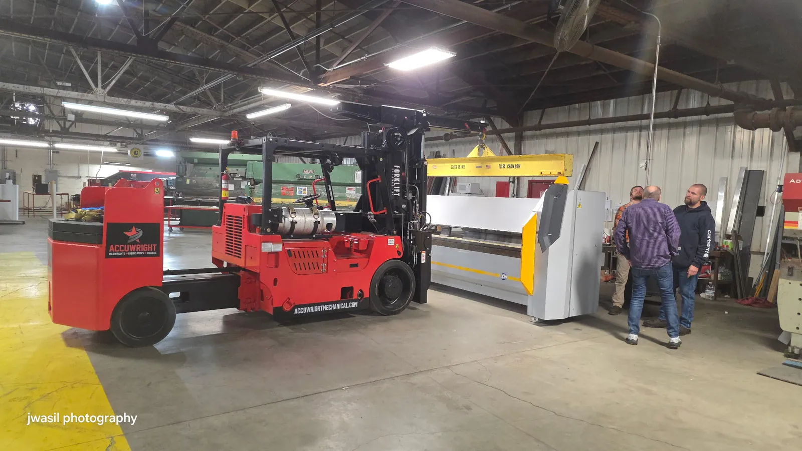 An Accuwright forklift lifting a machine on a yellow spreader beam while plant staff watch the pick