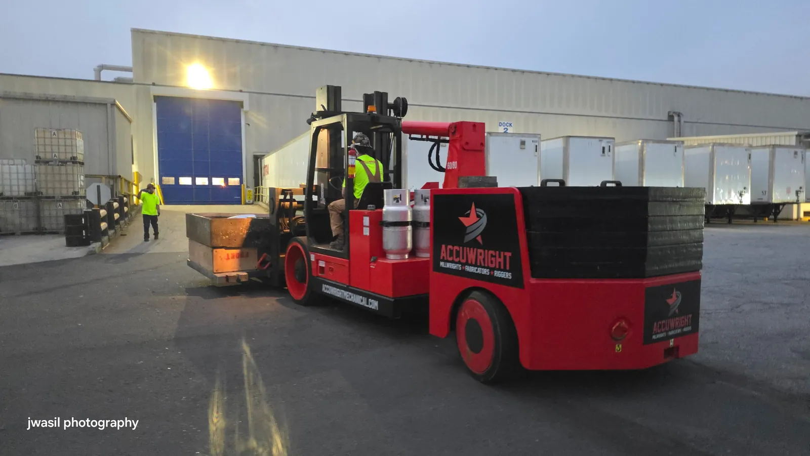 An Accuwright machinery mover carrying a crated load at a plant dock at dusk with a spotter ahead