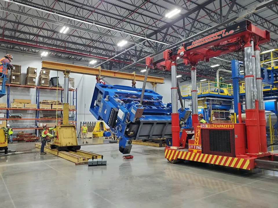 An Accuwright hydraulic gantry system lifting a large blue machine inside a bright plant while crew in hard hats and hi-vis spot the lift